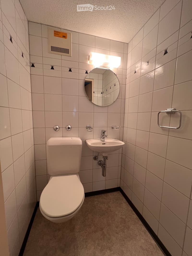 Modern bathroom with white tiled walls, a toilet, a sink with a modern faucet, a round mirror with built-in lighting, and a towel ring.