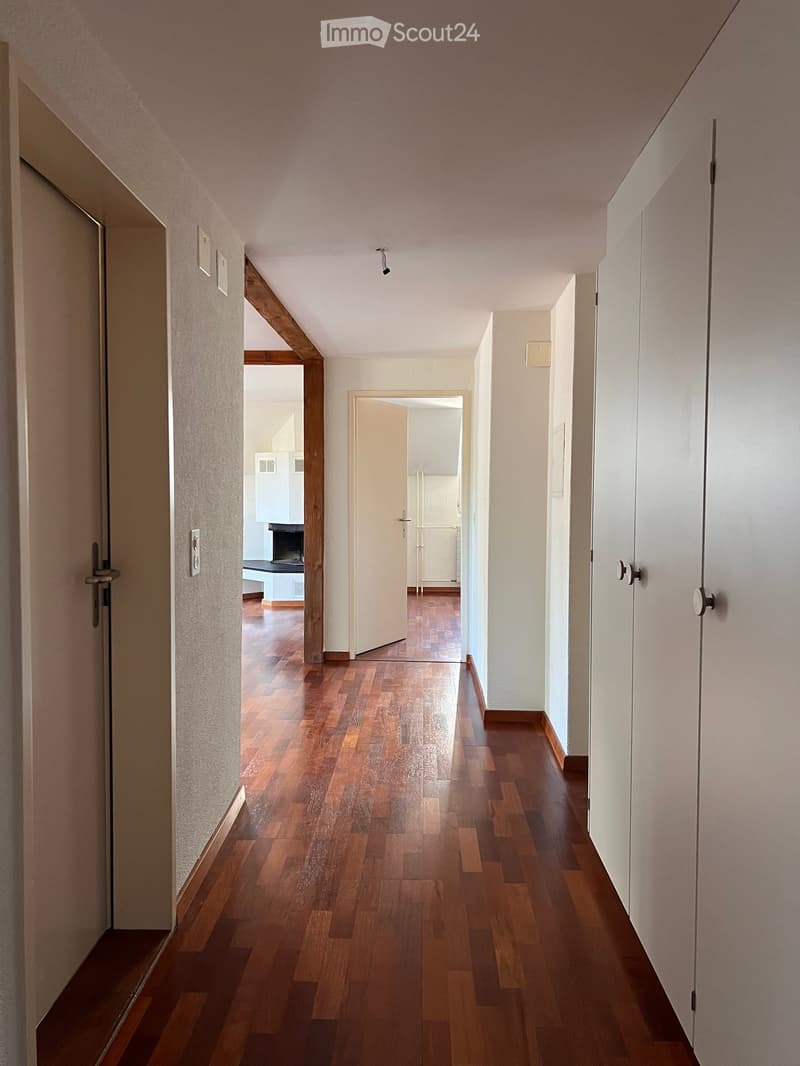 A long, narrow hallway with wooden flooring and white walls, featuring several doors and light switches.