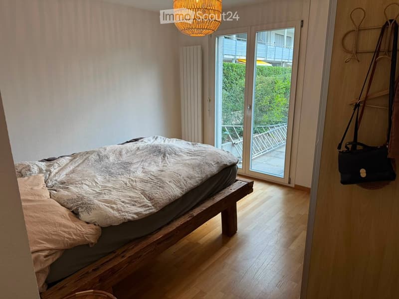 Cozy bedroom with wooden flooring, a bed with a textured duvet, a radiator, and a handbag hanging on the wall. Large windows offer a view of greenery and a balcony.