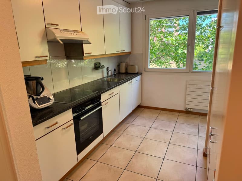 Modern kitchen with white cabinets, black countertop, tiled floor, and large windows overlooking greenery.