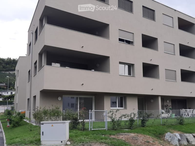 Modern multi-story residential building with balconies, large windows, and a fenced garden area with greenery and outdoor furniture.