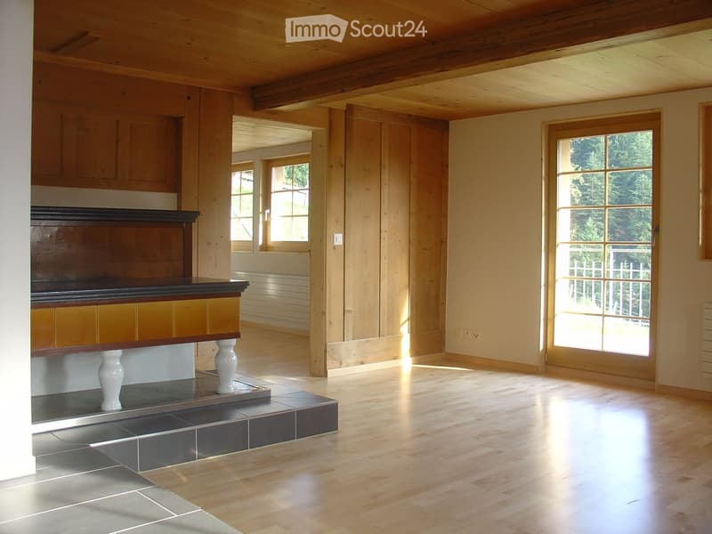 Bright, empty room with wooden walls, ceiling beams, and hardwood flooring. Features large windows and a raised platform with a dark wooden railing.