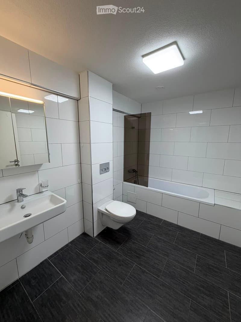 Modern bathroom with white tiles, dark grey floor, sink, toilet, bathtub, and a mirror with built-in lighting.