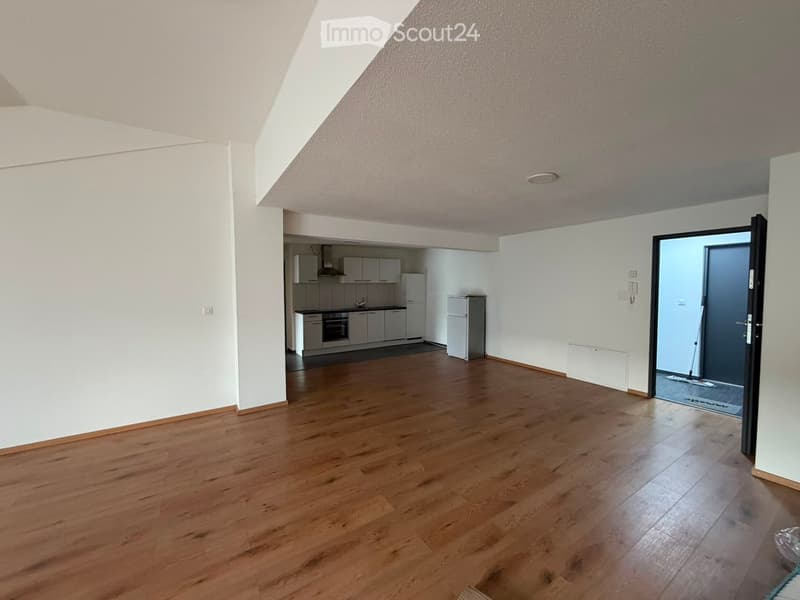 Empty room with wooden flooring, white walls, and a small kitchen area visible in the background. No furniture or personal items are present.