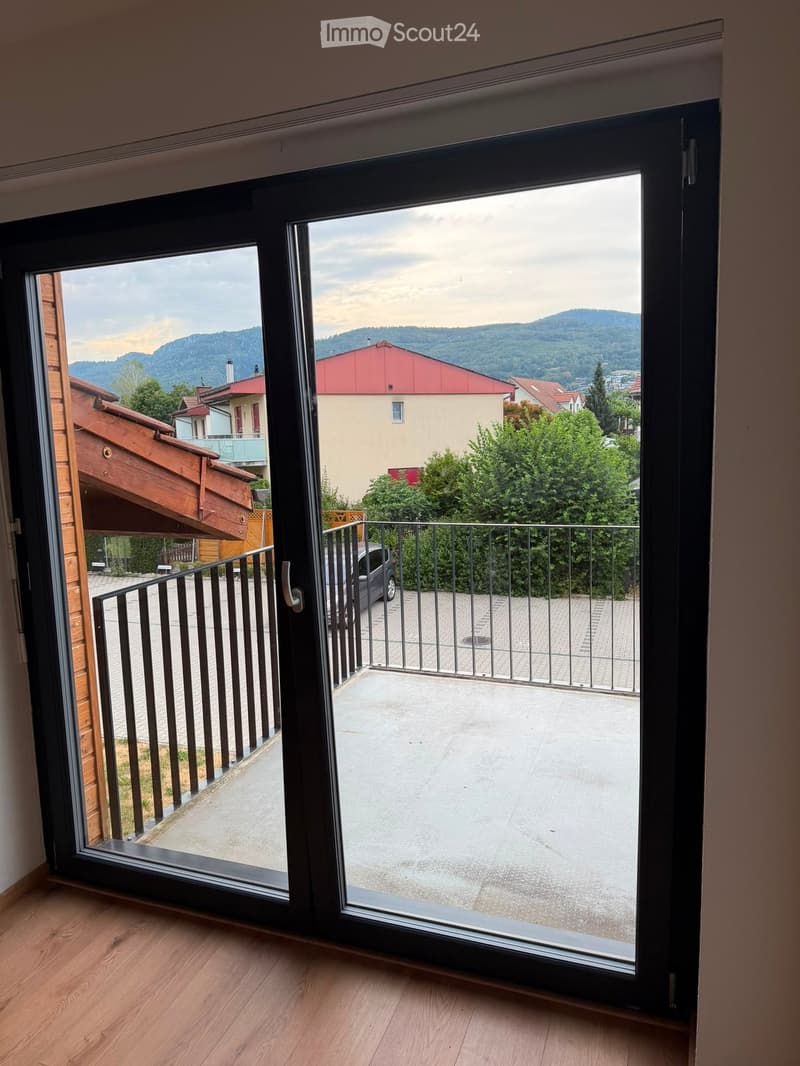 Open sliding balcony door with metal railing, overlooking a parking area and residential neighborhood with mountains in the background.