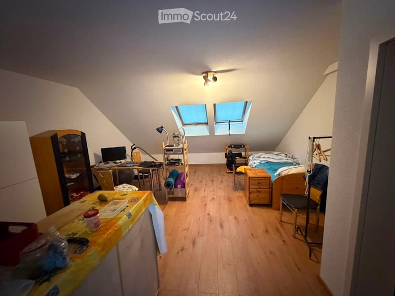 Cozy attic bedroom with wooden floors, slanted ceilings, and skylight windows. Features a bed, desk with laptop, chair, fan, and storage cabinet.