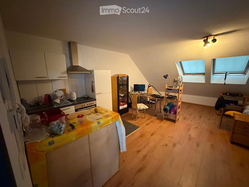 A combined kitchen and living space with wooden flooring, white cabinets, a small table with a yellow tablecloth, a desk with a computer, and a fan. The room features skylight windows and a slanted ceiling typical of an attic space.