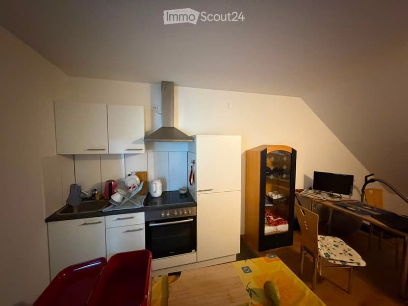A compact studio apartment with a kitchen area featuring an oven, sink, and fridge, and a living area with a chair, table, and laptop on a desk.