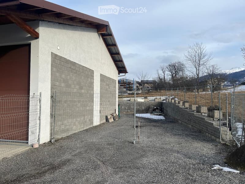 Modern concrete building with a garage entrance, surrounded by a gravel area and a fenced perimeter with a stone wall and small garden plots.