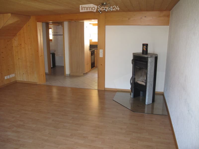 Bright, wooden-floored room with a modern fireplace and a small kitchen area visible through an open doorway.