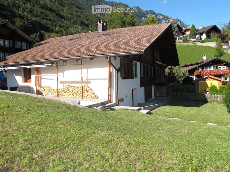 Traditional Swiss house with a tiled roof, wooden balcony, and a stack of firewood near the entrance, surrounded by a grassy lawn and neighboring houses in a mountainous area.