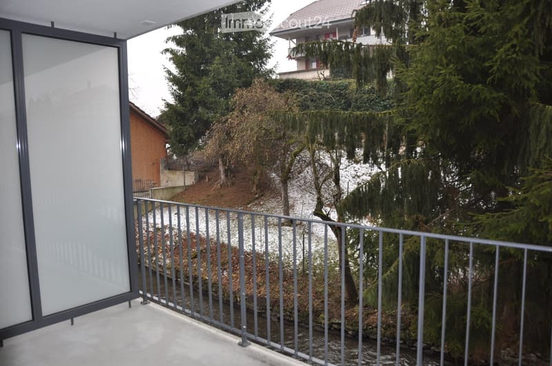 Balcony with metal railing, overlooking a landscaped garden area with trees and a neighboring house in the background.