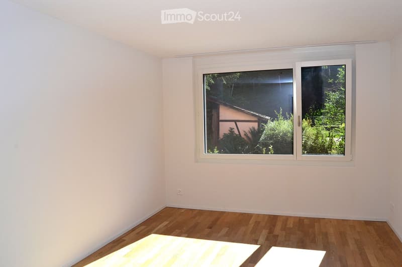 Empty room with light-colored walls, wooden flooring, and a large window showing greenery outside.