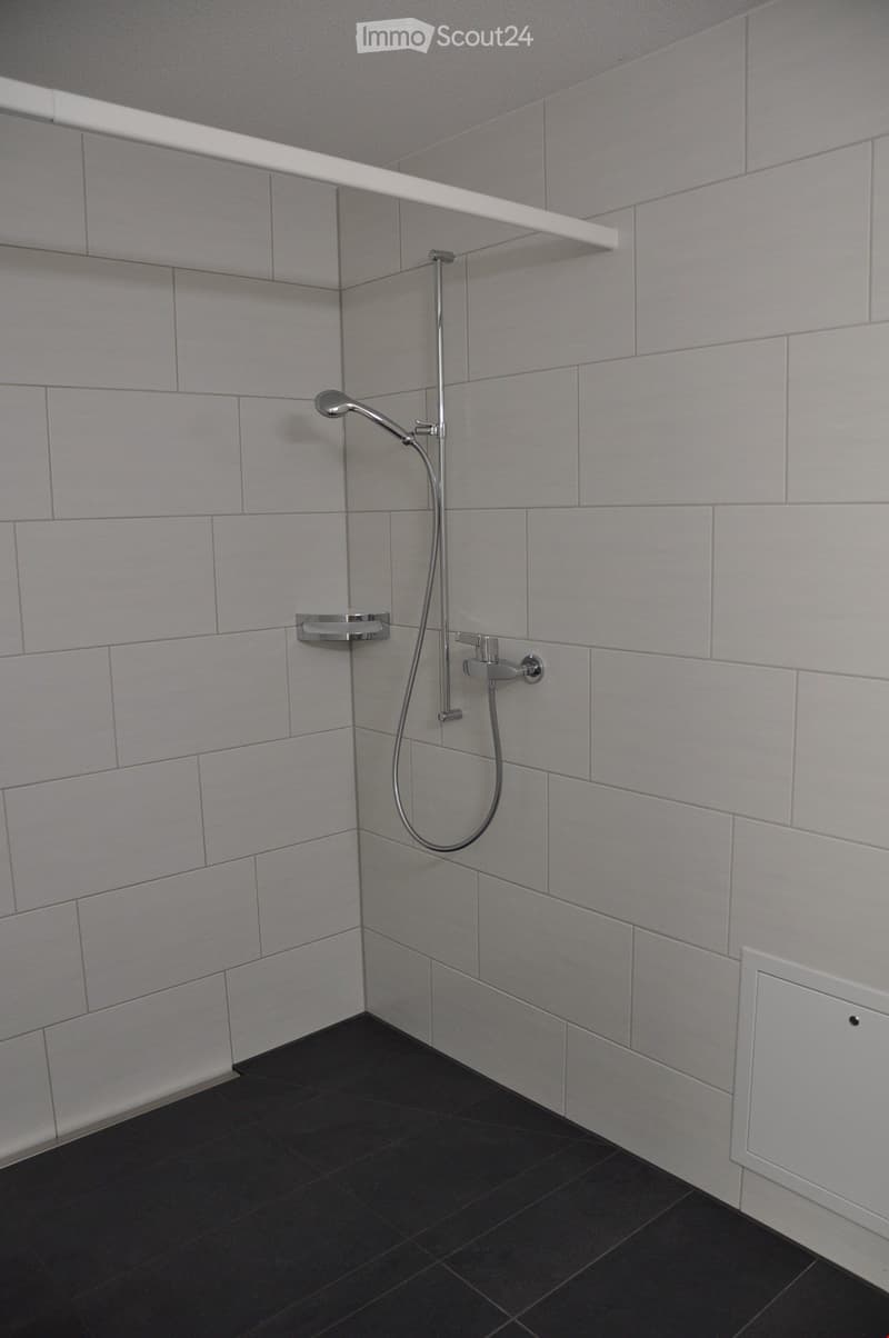 Modern shower area with grey tiles, black floor, chrome showerhead, faucet, and a white shelf above.