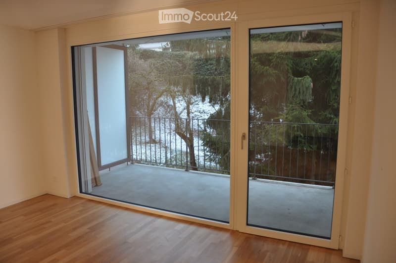 Sliding glass doors open to a balcony with a metal railing, overlooking a garden with trees.