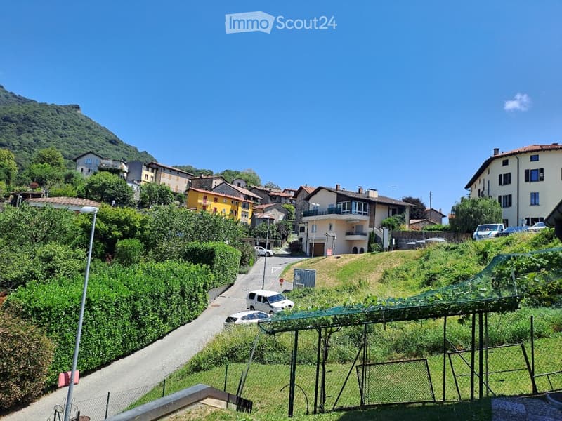 A scenic view of a hillside village with houses, greenery, and a winding road, surrounded by lush vegetation and a clear blue sky.