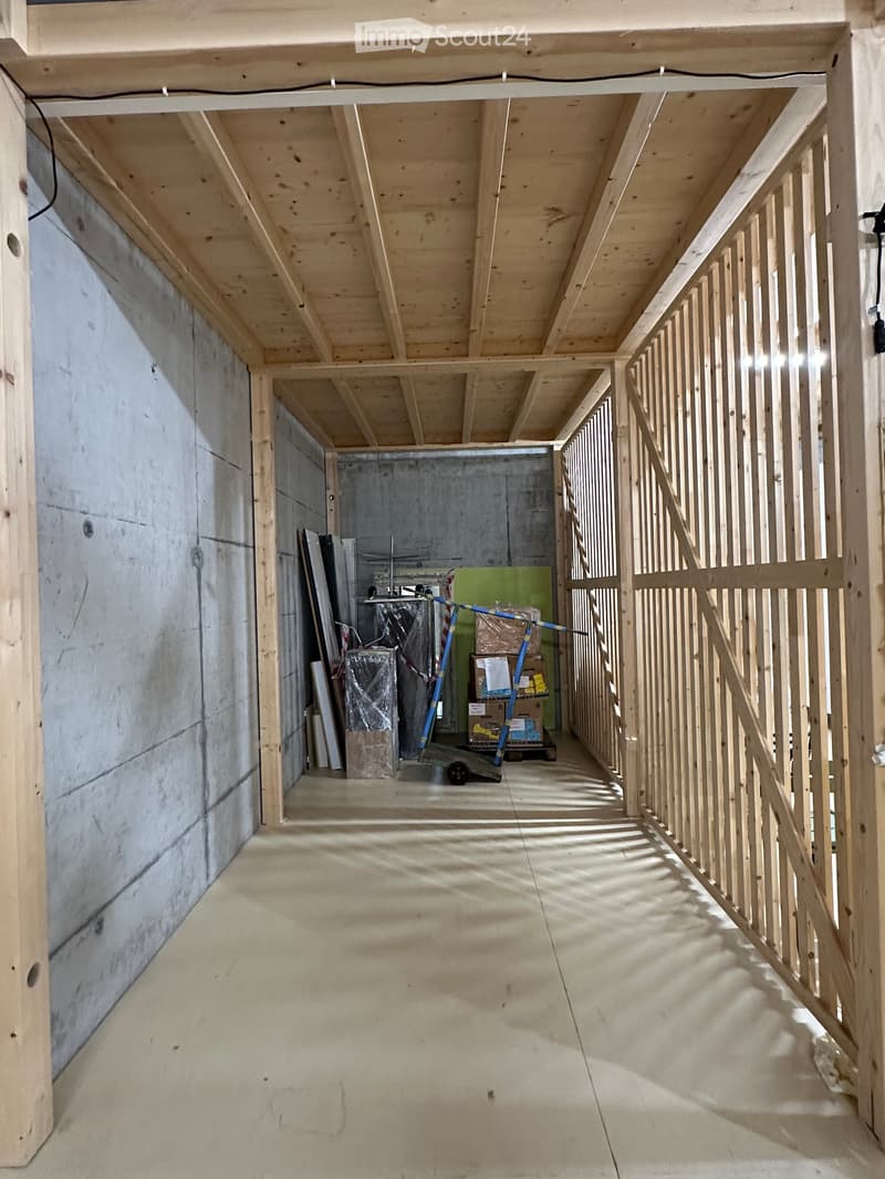 Empty unfinished hallway with exposed wooden beams and a staircase, concrete walls, and scattered construction materials in the background.