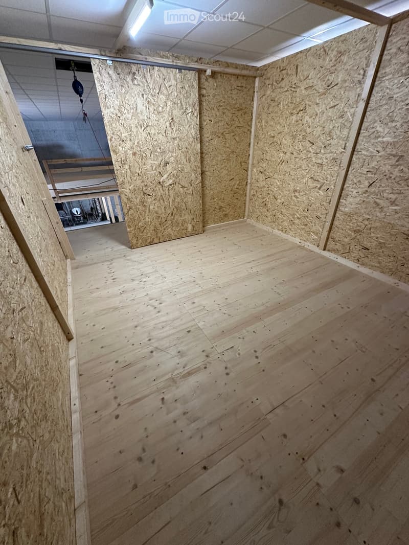 Empty room with wooden walls and floor, exposed beams, and a partially open doorway leading to another area.