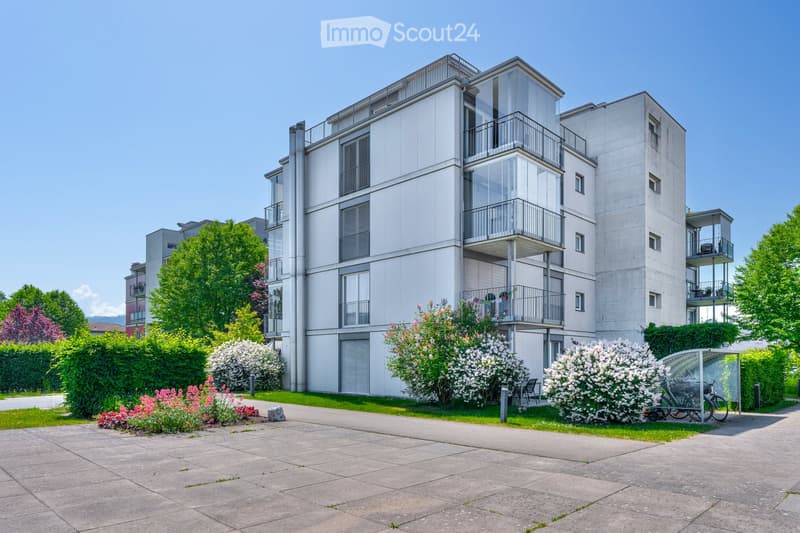 Modern multi-story residential building with balconies, surrounded by greenery and a paved walkway.