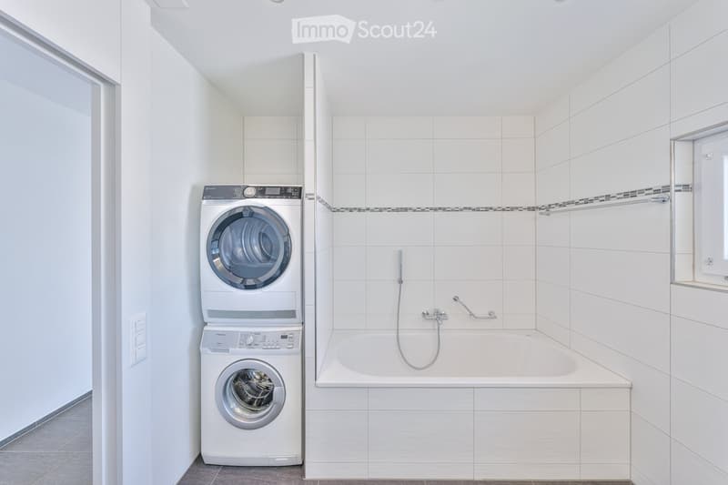 Modern bathroom with white tiled walls, a bathtub, and stacked washing machine and dryer unit.
