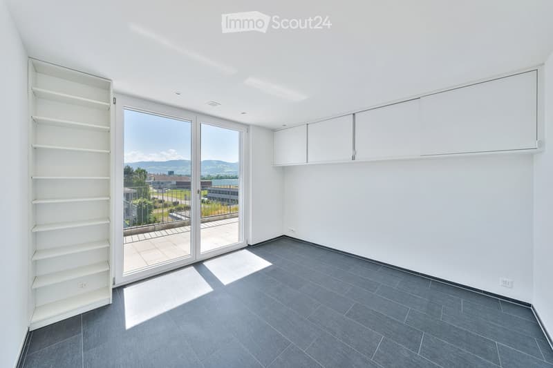 Empty room with white walls, grey tiled floor, built-in shelving unit, and large glass sliding doors leading to a balcony with a scenic view.