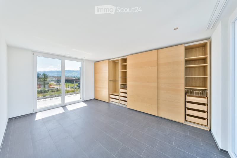Empty room with grey tiled floor, white walls, large sliding glass doors leading to a balcony, and built-in wooden closet with shelves and drawers.