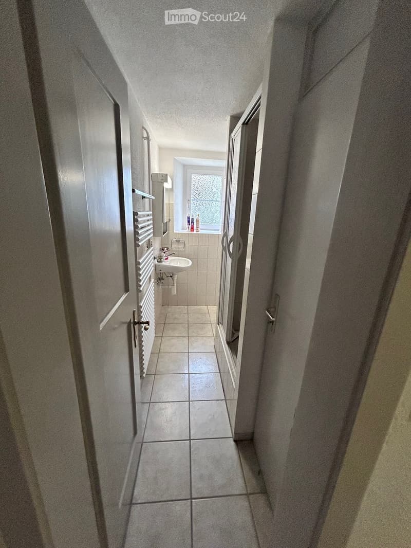 A narrow hallway with tiled flooring and light-colored walls, leading to a small bathroom with a sink, mirror, and window.