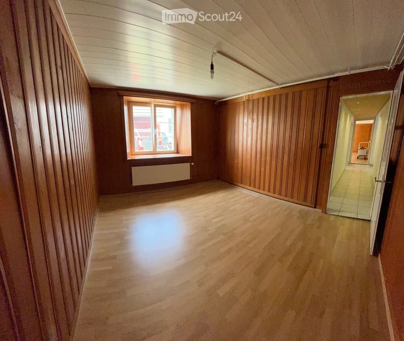 Empty room with wooden walls and floor, large window with radiator below, and a doorway leading to another area.