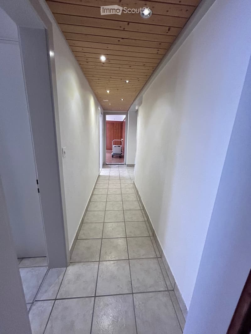 Clean, narrow hallway with tiled floor, white walls, and wooden ceiling with recessed lighting. A small cart is visible at the end of the hallway.