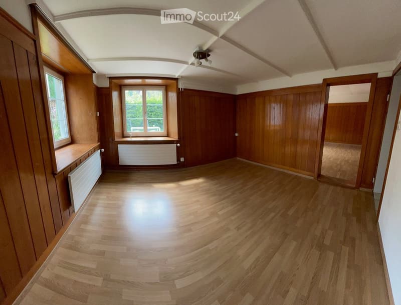 Empty room with wooden flooring, wooden panel walls, large windows, radiators, and a ceiling light fixture.