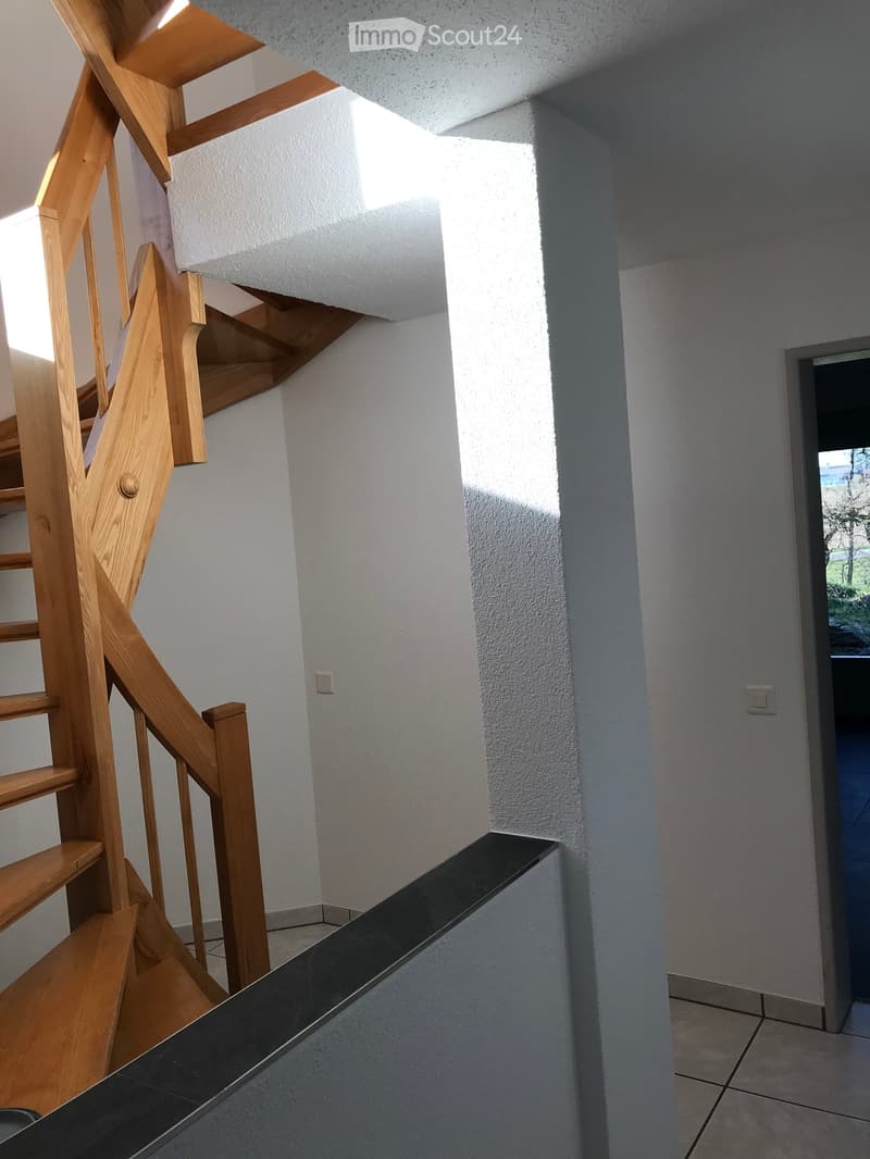 Modern hallway with wooden staircase, white walls, and tiled floor. Natural light streams in from the left side.