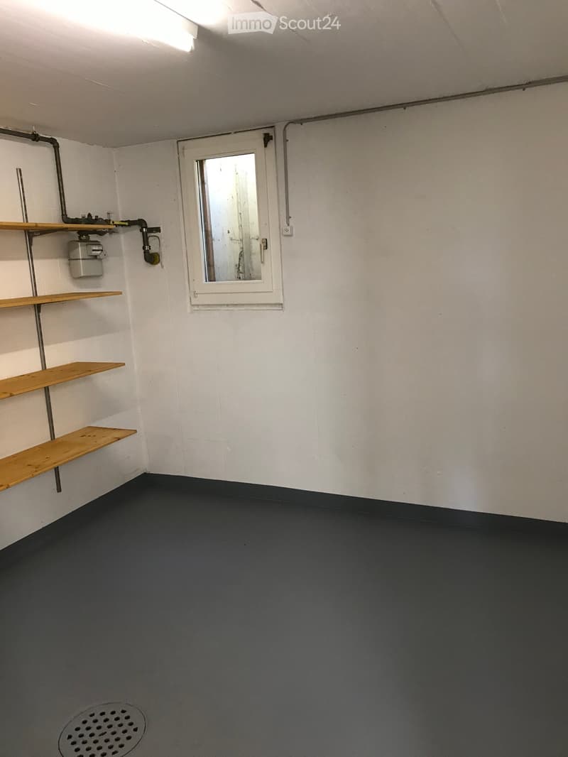 Empty room with white walls, dark grey floor, wooden shelves, a small window, and a drain cover on the floor.