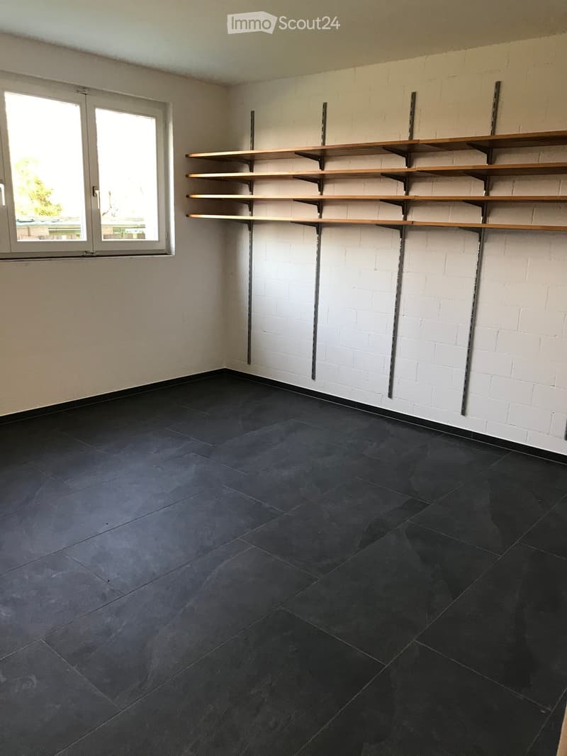 Empty room with dark grey tiled floor, white brick walls, and built-in wooden shelves on one wall. A large window with white frame is visible on the left side.
