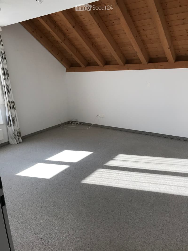 Empty room with grey carpeted floor, white walls, and wooden slanted ceiling beams. Sunlight streams in from a window on the left side.