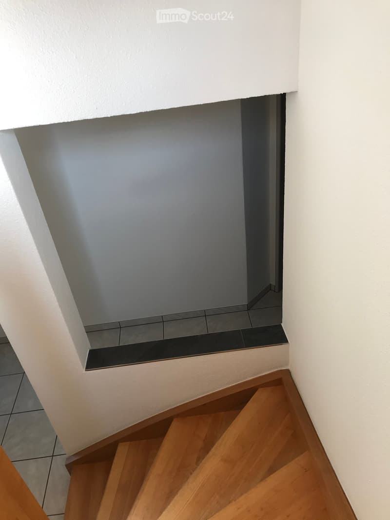 A modern staircase leading upwards with wooden steps and white walls, featuring a small landing area with a black-tiled floor and a white ceiling.