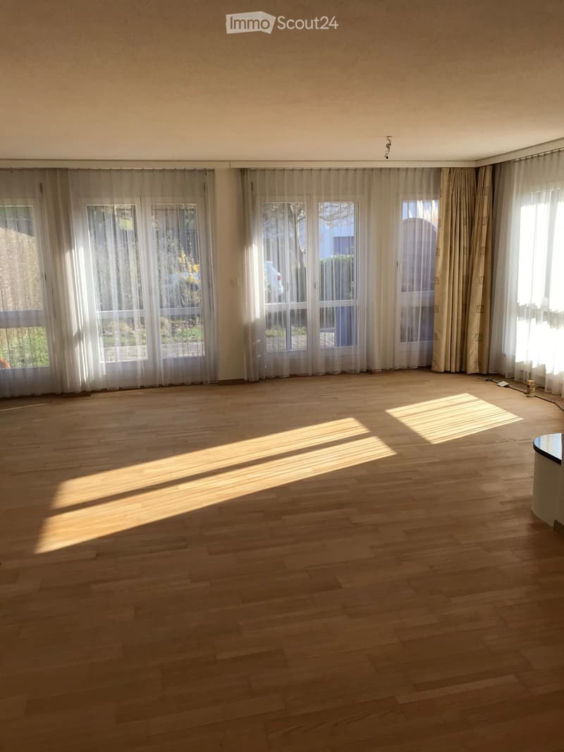 Empty room with wooden flooring, large windows with sheer curtains, and natural light streaming in.