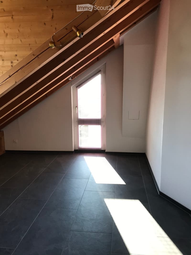 Empty attic space with dark tiled floor, wooden slanted ceiling, and a small window letting in natural light.