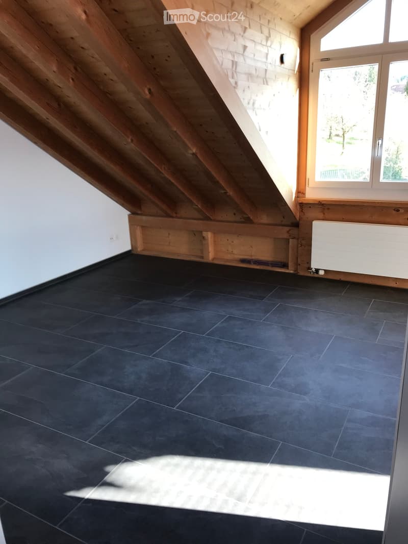 Empty attic room with dark grey tiled floor, wooden beams, white walls, and a window with wooden frame. No furniture or equipment present.