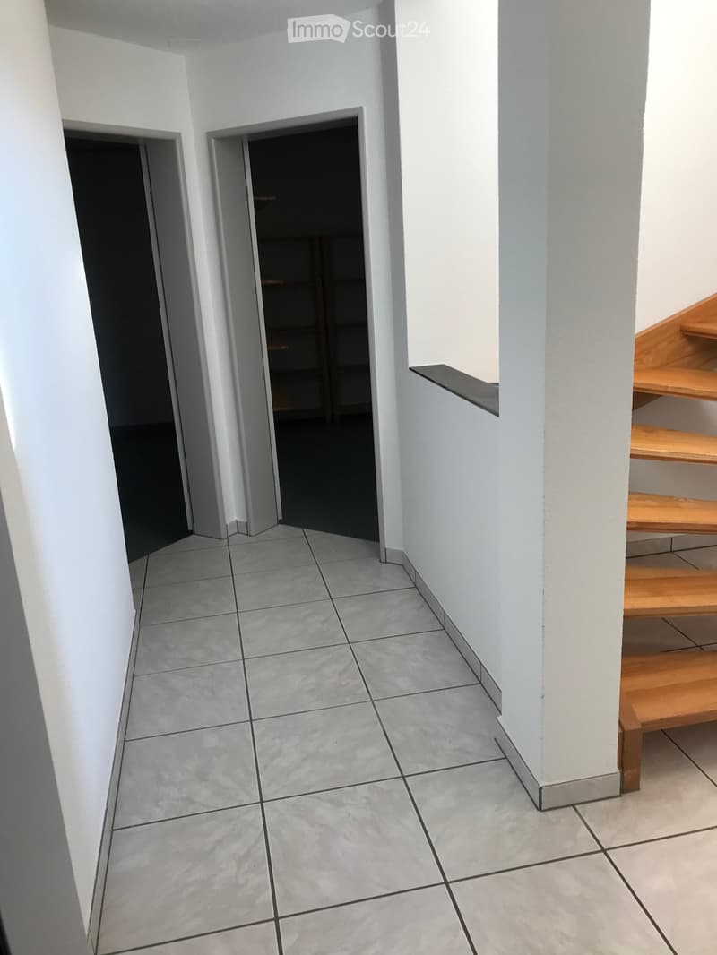 Empty hallway with tiled floor, white walls, and wooden staircase visible to the right.