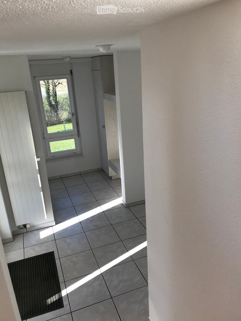 Empty hallway with tiled floor, white walls, and a door leading outside. Natural light streams in from the door window.