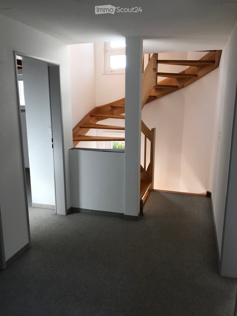 Empty hallway with carpeted floor, white walls, and a wooden staircase leading upwards.