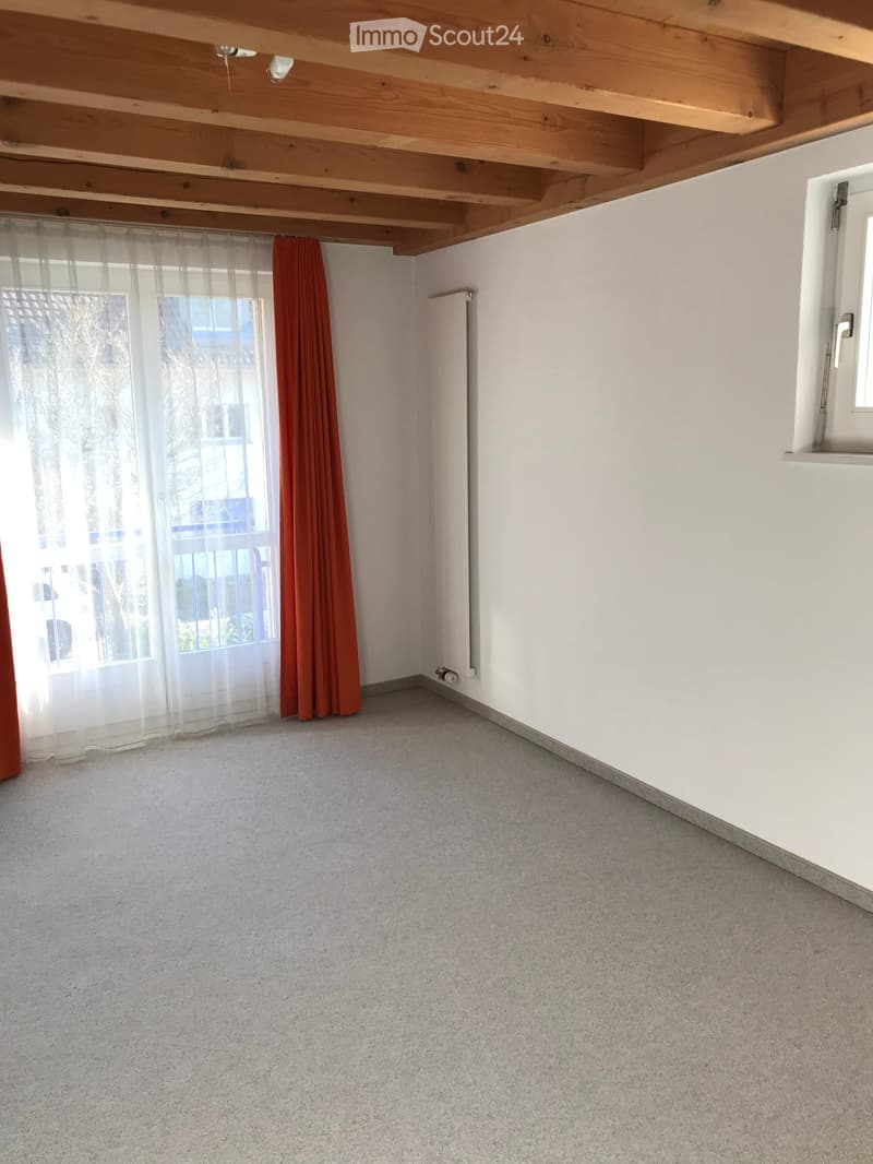 Empty room with light-colored walls, carpeted floor, and large windows with sheer and solid curtains. Wooden ceiling beams visible.