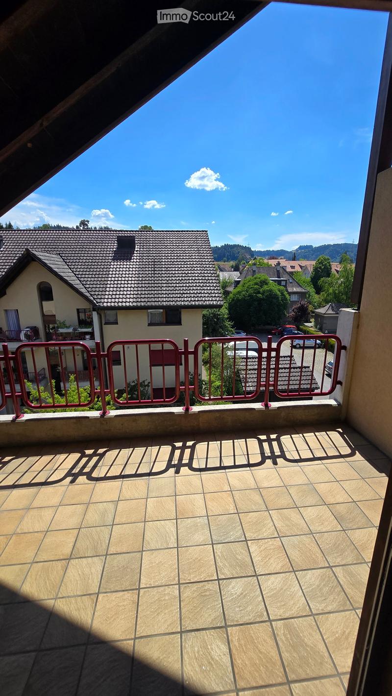 Balcony with tiled floor and red metal railing, overlooking a residential area with houses, trees, and parked cars.