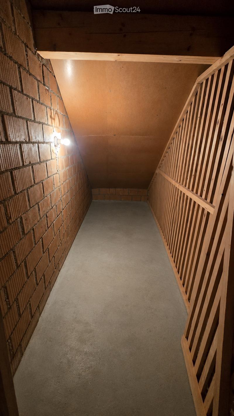A narrow, concrete-floored hallway with exposed brick walls and a wooden handrail on one side. The ceiling is slanted, and the lighting is minimal, coming from a single wall-mounted fixture.