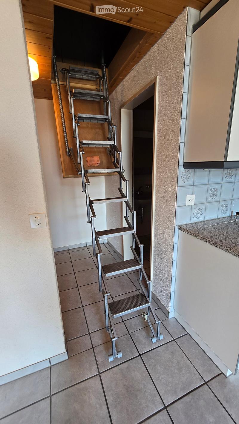 A narrow hallway with tiled flooring, a metal staircase leading upwards, and a partially visible kitchen area with white cabinets and a granite countertop.