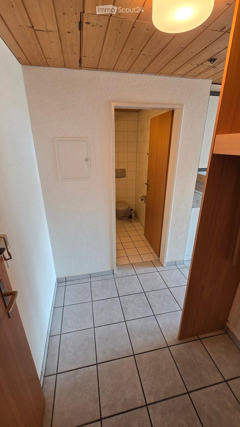 A narrow hallway with tiled flooring and light-colored walls, leading to a small bathroom with a toilet and tiled walls.