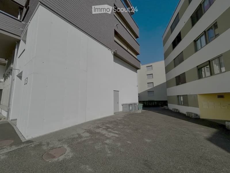 Modern multi-story residential building with white and grey exterior, numbered '13', featuring balconies and a paved ground area.