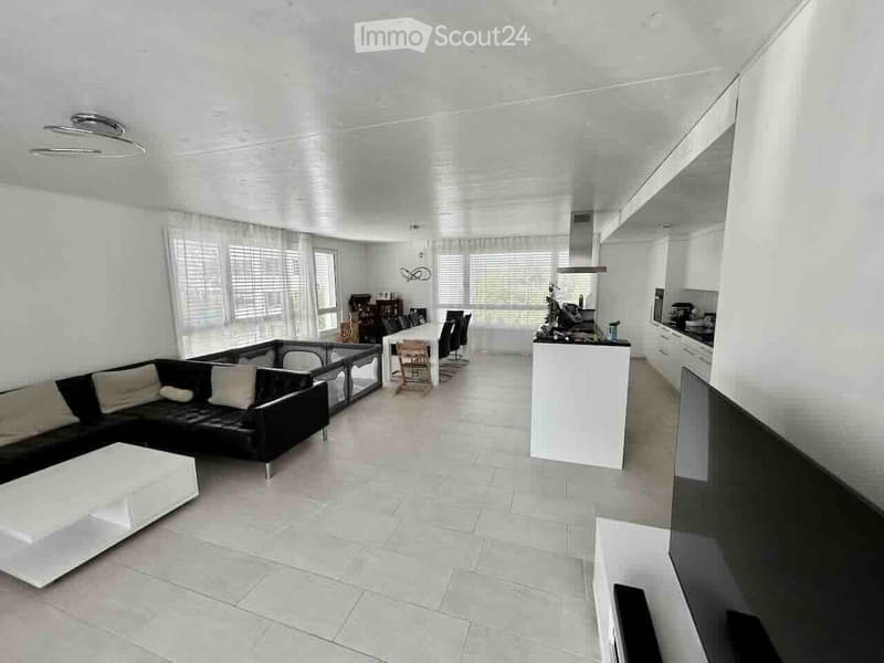 Open-plan living and dining area with a black sectional sofa, white coffee table, dining table with chairs, and a modern kitchen island with cabinets and appliances.