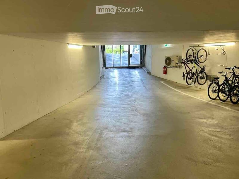 Underground parking corridor with concrete floors, white walls, and bicycles stored on racks. Natural light enters through a glass door at the far end.