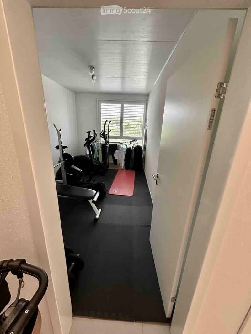 A small, well-lit exercise room with a black rubber floor, containing an exercise bike, treadmill, elliptical machine, yoga mat, and a clothing rack with hanging clothes.